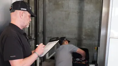 Ein Service-Techniker arbeitet im Aufzug, der andere steht davor und notiert etwas auf einem Blatt Papier.