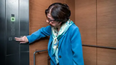 An elderly lady wearing a blue jacket and glasses stands in an elevator and presses the emergency button.