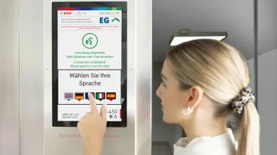 Eine Frau steht in einem Aufzug und tippt mit ihrer Hand auf das Display des Zwei-Sinne-Notrufs.