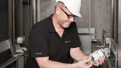 Detlef se sienta en el hueco de un ascensor y mira un instrumento de medición. Lleva un casco de seguridad.